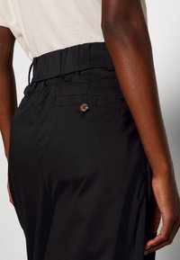Svarta byxor med elastisk midja, sidficka och en liten brun knappdetalj, med en slät tygtextur och avslappnad passform.
