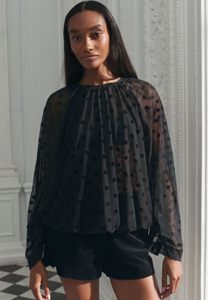 Ženska z dolgimi temnimi lasmi, oblečena v prosojno črno bluzo z belimi pikami in balonastimi rokavi ter črne kratke hlače, stoji v svetli sobi z okrašeno štukaturo.