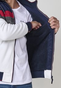Pánska bunda na zips, ktorá má bielu vonkajšiu látku a námornícky modrý interiér, textúrovanú podšívku a malú vnútornú kapsu na uskladnenie.