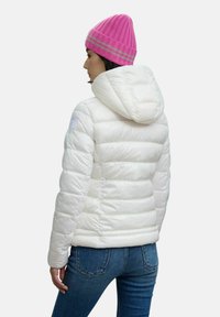 Witte gewatteerde jas met een capuchon, voorzien van een gequiltde textuur en een logo op de schouder. Gecombineerd met blauwe jeans en een felroze ribbelgebreide muts.