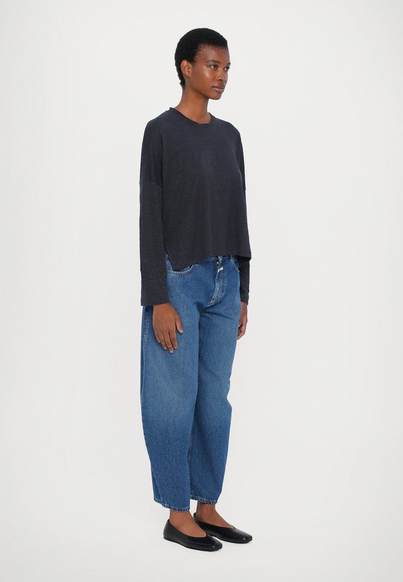 Lockere dunkelblaue Langarmbluse mit asymmetrischem Saum, kombiniert mit weiten blauen Jeans und schwarzen Ballerinas. Einfaches Design mit minimalen Akzenten.