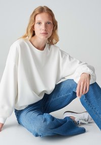 Felpa bianca oversize con spalle a caduta, colletto a costine e dettagli di polsini elasticizzati, abbinata a jeans azzurri chiari e scarpe da ginnastica bianche.