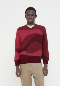 Μπορντό πλεκτό πουλόβερ με λαιμόκοψη σε σχήμα V, διακοσμημένο με κόκκινα κυματιστά μοτίβα σε αντίθεση και ριμπ μανσέτες και ρέλι. Κεντημένο λογότυπο στο κάτω μέρος.