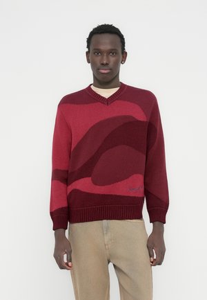 Μπορντό πλεκτό πουλόβερ με λαιμόκοψη σε σχήμα V, διακοσμημένο με κόκκινα κυματιστά μοτίβα σε αντίθεση και ριμ π μανσέτες και ρέλι. Κεντημένο λογότυπο στο κάτω μέρος.