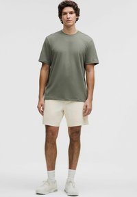 Tricou verde măsliniu dintr-un material moale, cu guler rotund și mâneci scurte; asortat cu pantaloni scurți crema și adidași deschisi, toate într-o paletă de culori neutră.