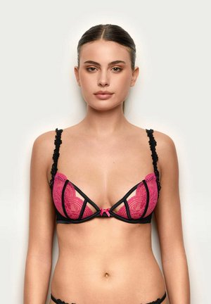 Kobieta ubrana w różowy koronkowy biustonosz z czarną lamówką i małą różową kokardką na środku, pozująca na jasnym, jednolitym tle.