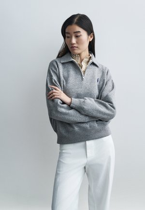 Ženska z dolgimi temnimi lasmi, oblečena v sivi pulover s četrtinskim zadrgo čez črtasto srajco in belimi hlačami, stoji z rokami prekrižanimi in gleda vstran.