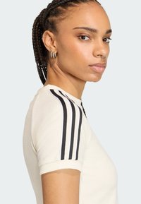 Krátky rukáv sveter v krémovej farbe s čiernymi pruhmi na pleciach, rebrovaným golierom a mäkkou textúrou, ktorý predvádza priliehavý strih.