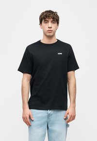 Νεαρός άνδρας με καστανά σγουρά μαλλιά, φορώντας μαύρο t-shirt με την επιγραφή «CONS» και ανοιχτό μπλε τζιν, στέκεται μπροστά σε απλό λευκό φόντο.