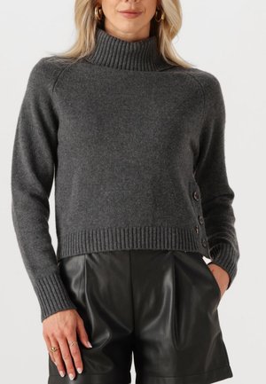 Donna che indossa un maglione a collo alto a coste di colore grigio scuro con bottoni laterali e pantaloni corti in pelle nera plissettati, mano in tasca, orecchini a cerchio dorati.