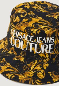 Čierny klobúk so širokým okrajom s zlatým kvetinovým vzorom, s nápisom "VERSACE JEANS COUTURE" v bielej farbe, vyrobený z odolného materiálu s štruktúrovaným povrchom.