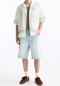 Hombre que lleva puestos unos pantalones cortos de mezclilla azul claro con un estampado, una camiseta de manga corta blanca con motivos de sol y naturaleza en verde, y zapatillas marrones con calcetines blancos.