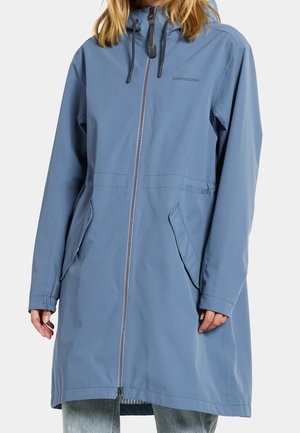 Femme portant un long imperméable bleu à capuche avec fermeture éclair frontale et poches en biais, associé à un jean bleu clair.