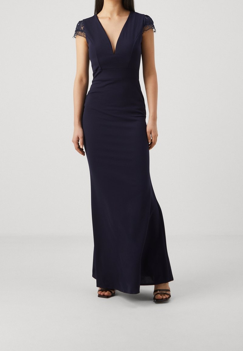 Femme portant une robe bleu marine longue jusqu'au sol avec des manches courtes en dentelle, un décolleté en V profond et des talons ouverts noirs sur un fond uni.