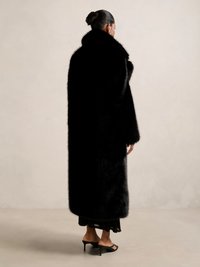 Svart pälsjacka med en lång, oversized design, hög krage och texturerad yta. Bärs över en svart klänning och sandaler.