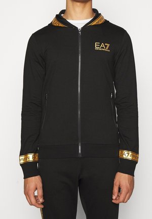 Mężczyzna w czarnej bluzie z kapturem na zamek i spodniach z złotym logo EA7 Emporio Armani na piersi, mankietach i krawędzi kaptura.