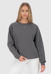 Felpa grigia con maniche raglan, scollo rotondo e polsini a costine. Il tessuto appare morbido e con una texture liscia. Abbinata a pantaloni bianchi.