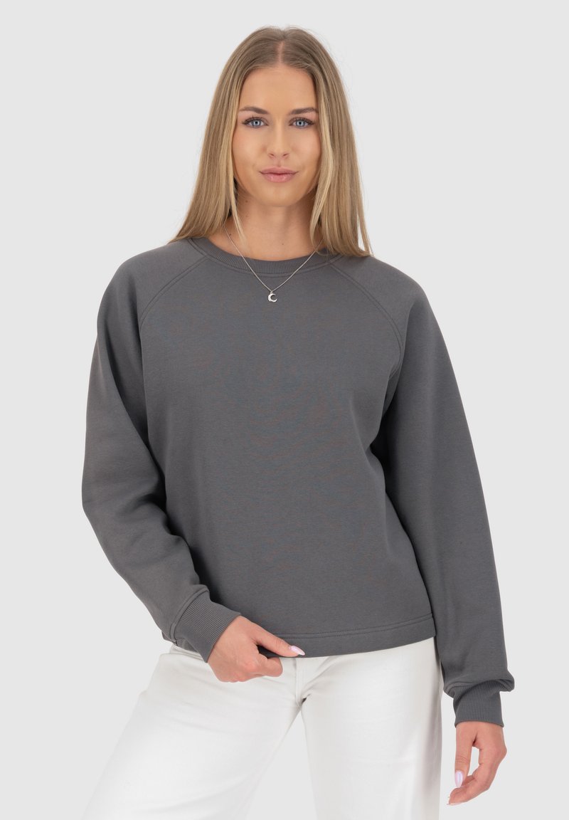Felpa grigia con maniche raglan, scollo rotondo e polsini a costine. Il tessuto appare morbido e con una texture liscia. Abbinata a pantaloni bianchi.