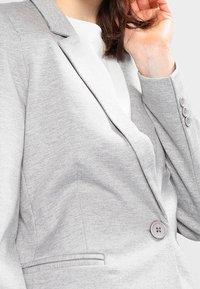 Šedý blejzr s klopovým límcem, jednobuttonovým zapínáním a jemnou strukturou. Obsahuje jednu kapsu a čtyři knoflíky na rukávu.