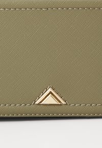 Tissu textured vert olive avec des ferrures triangulaires dorées dans le coin inférieur, présentant des détails de couture contrastés.