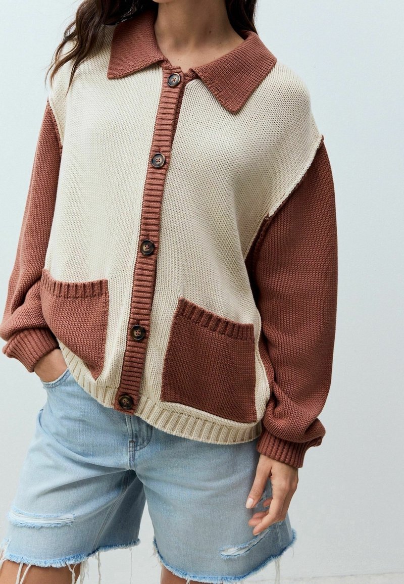 Femme portant un cardigan en tricot bicolore beige et marron boutonné avec des poches avant, associé à un short en jean bleu clair déchiré.