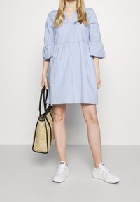 Hellblaues Kleid mit V-Ausschnitt, dreiviertellangen Ärmeln und einem mehrlagigen Design. Gestylt mit einer strukturierten beigen Tragetasche und weißen Sneakers.