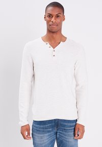 Helle beige Langarmshirt mit Henley-Ausschnitt, versehen mit drei braunen Knöpfen und einer weichen Textur. Kombiniert mit blauen Jeans.
