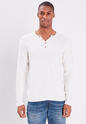 Lichtbeige langemouwen shirt met een henley-halslijn, voorzien van drie bruine knopen en een zachte structuur. Gekoeld met blauwe spijkerbroek.