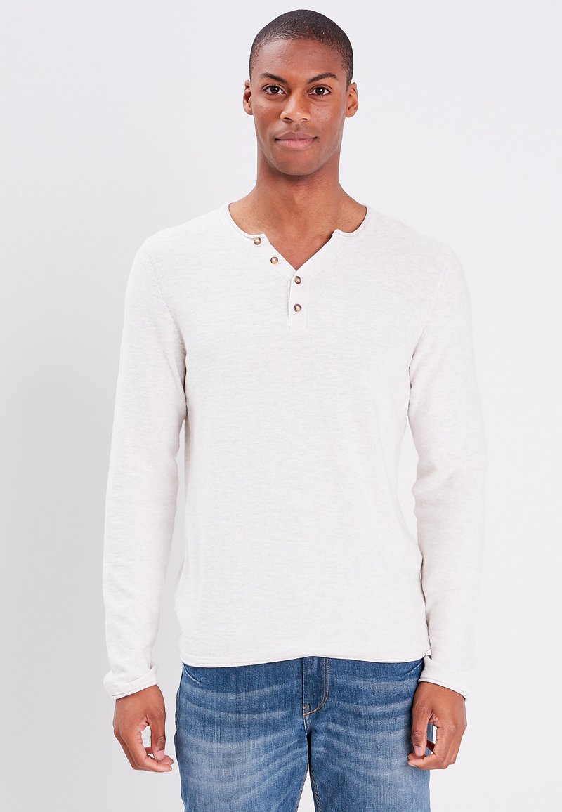Helle beige Langarmshirt mit Henley-Ausschnitt, versehen mit drei braunen Knöpfen und einer weichen Textur. Kombiniert mit blauen Jeans.