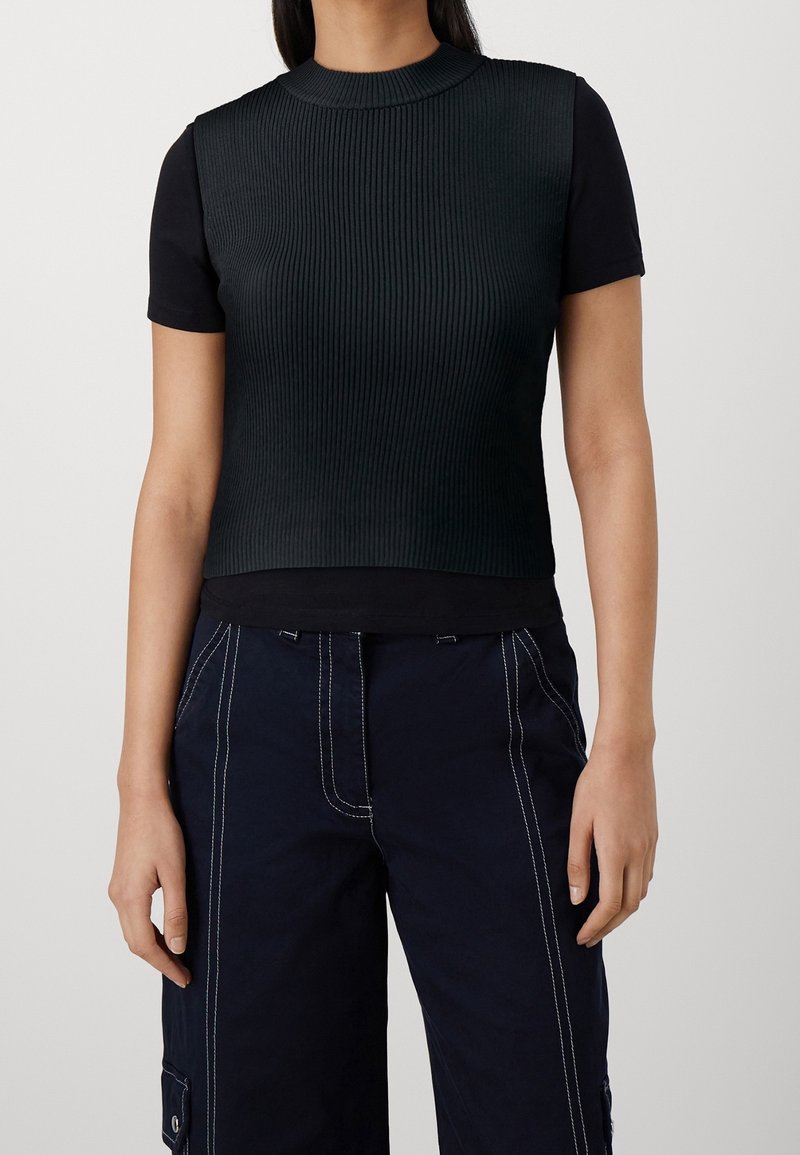 Femme portant un haut noir côtelé sans manches par-dessus un t-shirt noir à manches courtes et un pantalon foncé avec des détails de couture blancs.
