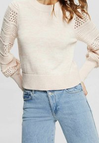 Pull en tricot rose clair avec des détails en dentelle sur les manches, des poignets côtelés et un ourlet ajusté, associé à un jean bleu clair.