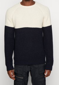 Pull en tricot avec une partie supérieure crème et une partie inférieure bleu marine, présentant un motif texturé et des poignets et ourlet côtelés. Coupe décontractée.