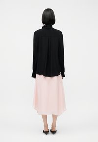Persona con cabello corto y oscuro, que lleva una blusa negra de manga larga y una falda midi rosa claro, de pie de espaldas sobre un fondo liso.