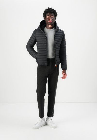 Veste matelassée noire avec capuche, design matelassé, portée sur une chemise gris clair et associée à un pantalon sombre et des baskets blanches.