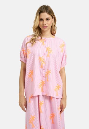 Young woman with long blonde hair wearing a pink and white striped button-up shirt and skirt set with orange palm tree prints.