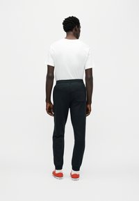 Μαύρο παντελόνι φόρμας με κορδόνι στη μέση και ελαστικές μανσέτες, συνδυασμένο με λευκό μπλουζάκι, εμφανίζεται από πίσω. Φαίνονται κόκκινα αθλητικά παπούτσια.