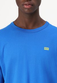 Μπλε βαμβακερή μπλούζα με στρογγυλή λαιμόκοψη. Διαθέτει ένα μικρό κίτρινο λογότυπο "DIESEL" στην περιοχή του στήθους. Λαμπερή υφή, χαλαρός σχεδιασμός.