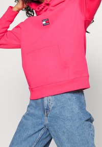 Persona che indossa un luminoso hoodie rosa di Tommy Jeans e jeans azzurri, in posa con le braccia alzate contro uno sfondo neutro.