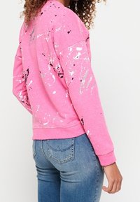 Rosa sweatshirt med stänkmålningsdesign i vitt och svart. Material av bomull, rund halsringning, ribbade muddar och fåll. Kombinerad med blå jeans.