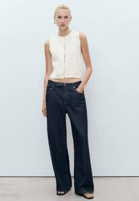 Weißes ärmelloses Top mit vorderen Knöpfen, kombiniert mit dunkelblauen weiten Jeans. Minimalistisches Design, glatte Textur und kontrastierende Farben.