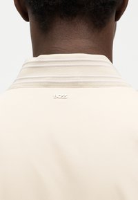 Vue arrière d'une personne portant une veste crème avec un col côtelé et un petit logo blanc "BOSS" sur le haut du dos.