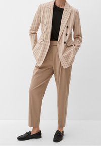 Blazer beige a righe sottili con bottoni neri, abbinato a pantaloni chiari beige su misura e mocassini neri, creando un look moderno e coerente.