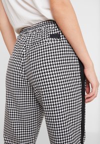 Kalhoty s potiskem houndstooth v černobílé barvě s černým elastickým pasem a postranním pruhem, obsahující zadní kapsu a texturovanou tkaninu.