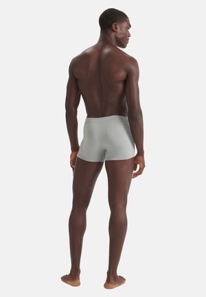Man staat op blote voeten met de rug naar de camera, gekleed in lichtgrijze strakke boxershorts tegen een effen witte achtergrond.