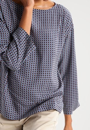 Femme portant un chemisier à manches longues à motifs avec des formes géométriques bleues, blanches et noires, associé à un pantalon beige clair.