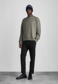 Olivgrüner Pullover mit Rundhalsausschnitt, schwarze Hose und schwarze Loafers. Gestylt mit einer marineblauen Strickmütze. Einfaches, modernes Design.