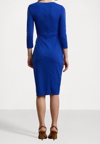 Blå, figurnära klänning med trekvartsärmar och rund halsringning. Har dragkedja baktill och knälång fåll, kombinerad med bruna klackskor.