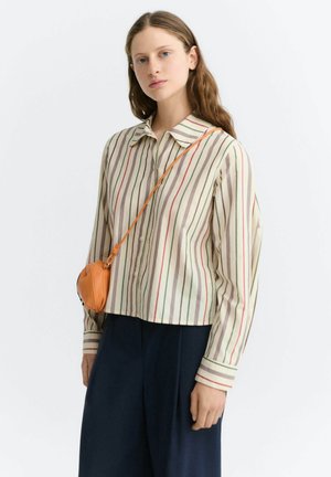 Jeune femme aux longs cheveux portant une chemise rayée verticale multicolore et un pantalon foncé avec un sac orange porté en bandoulière, debout sur fond blanc.