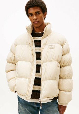 Joven con una chaqueta acolchada beige sobre un suéter de rayas marrón y blanco y jeans azules frente a un fondo liso.