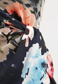 Detailní záběr na látku s texturovaným květinovým vzorem v námořnické modři, broskvové, světle modré a černé barvě, s ozdobným zkrouceným uzlem.